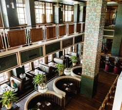 Interior of a stylish, two‑level restaurant with dark wood booths, green brick walls, and tall windows, plus a floral-patterned column centerpiece.