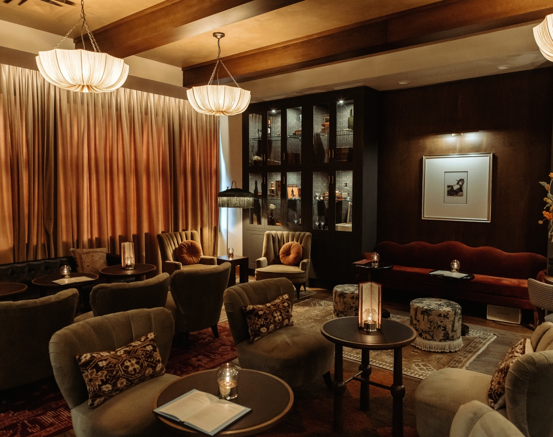 Luxurious hotel lounge with tufted armchairs, round tables, and warm ambient lighting under wooden beams with curtains.