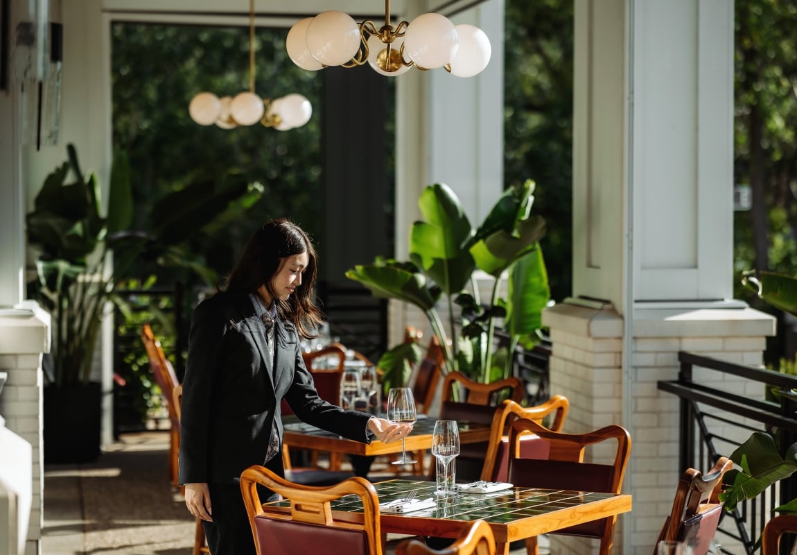 Woman in a black blazer stands at an outdoor restaurant table with a wine glass in hand.