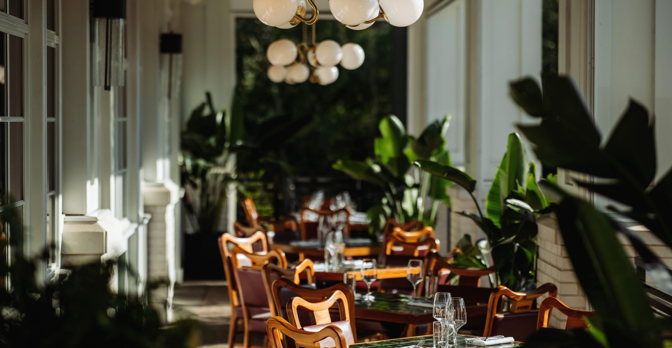 Sunlit restaurant terrace with wooden chairs, tiled tables set with glassware, and round globe lights hanging from the ceiling, flanked by green plants.