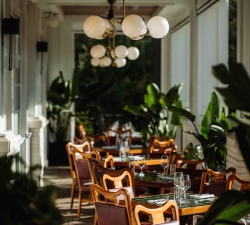 Sunlit restaurant terrace with wooden chairs, tiled tables set with glassware, and round globe lights hanging from the ceiling, flanked by green plants.