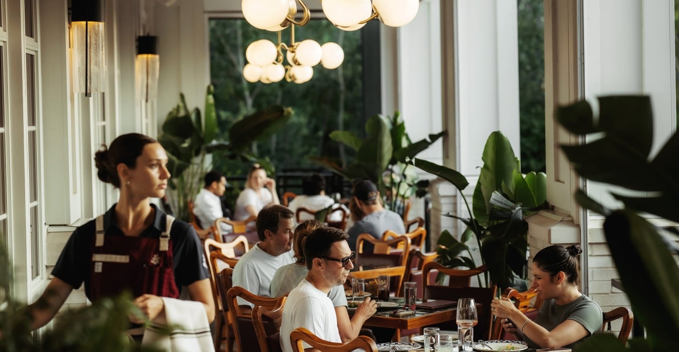 People dining in a sunlit cafe along a windowed wall with wooden chairs and green plants, a globe chandelier overhead
