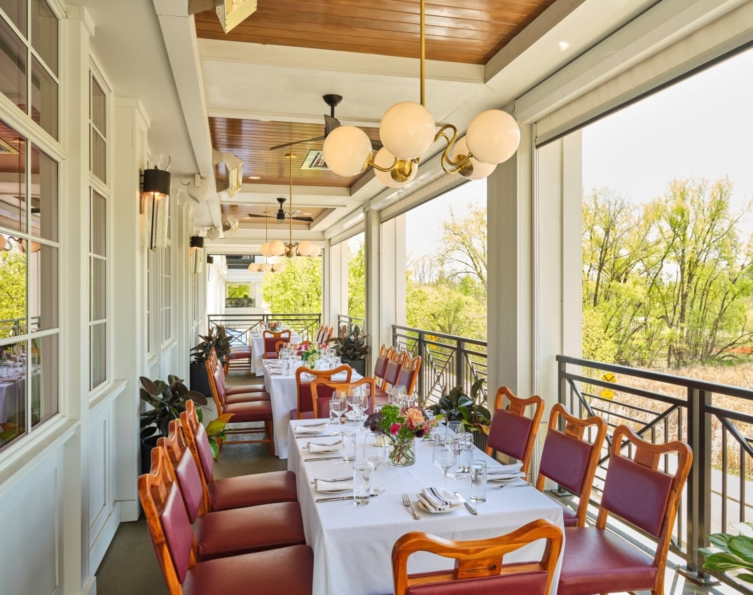 Outdoor restaurant balcony with long white-tablecloth tables set for dinner, wooden chairs, glassware, and greenery outside the railing, bright daylight.