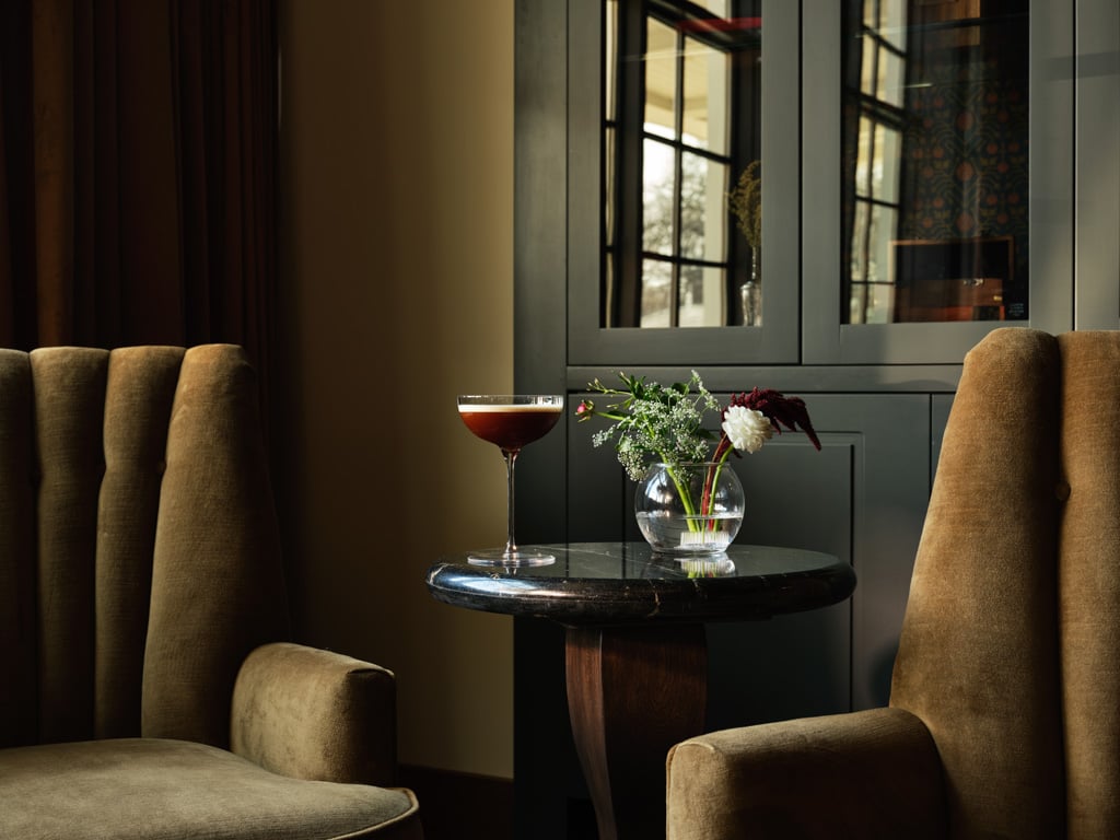 Cocktail glass on a round dark table between two beige-brown chairs in a cozy lounge with a vase of flowers nearby.