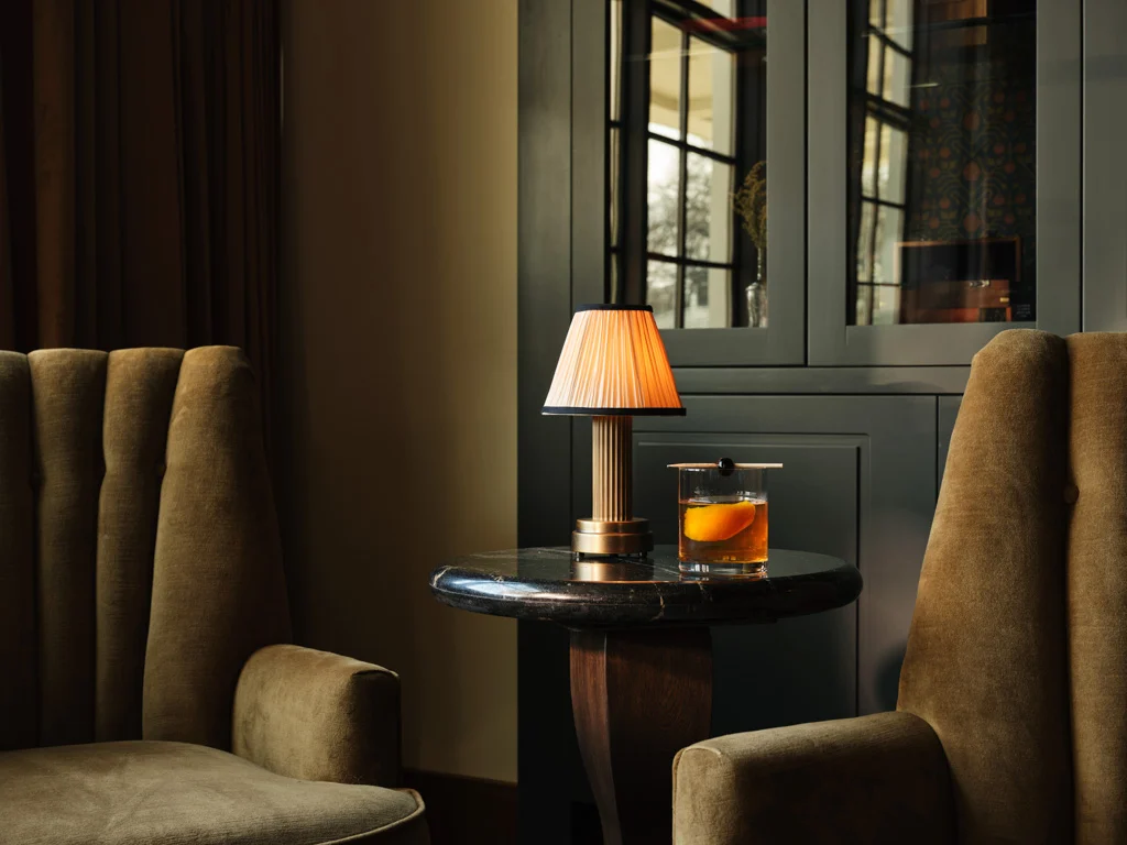 Cozy lounge scene with a lit table lamp and a glass of whiskey on a round marble table between two tan upholstered chairs.