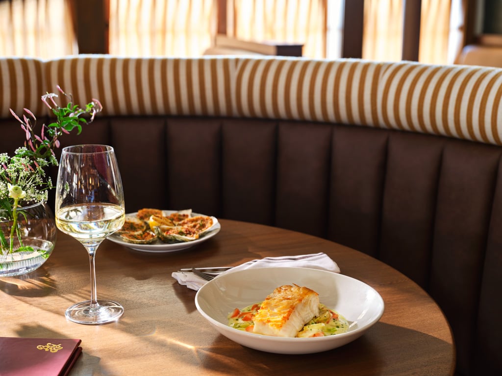 Table at a restaurant with a glass of white wine, a plated fish dish, and a small bouquet of flowers on the table.