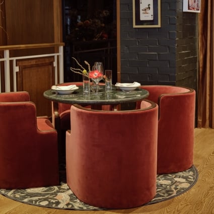 Round marble table set for four with red velvet chairs in a dimly lit restaurant corner on a patterned rug, with a flower arrangement nearby.