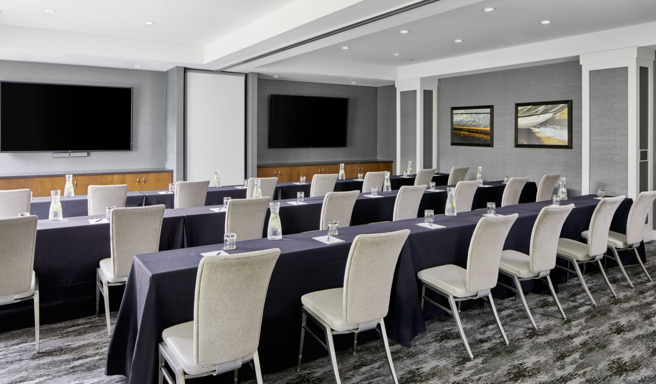 Conference room arranged for a meeting with long black-tablecloth tables, beige chairs, water bottles and glasses, and two wall-mounted TVs on gray walls.