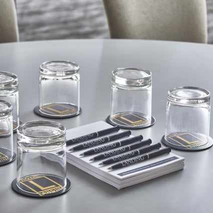 Table setup with several inverted glasses on round branded coasters, a stack of LANDING pens, and a carafe with lime slices nearby.