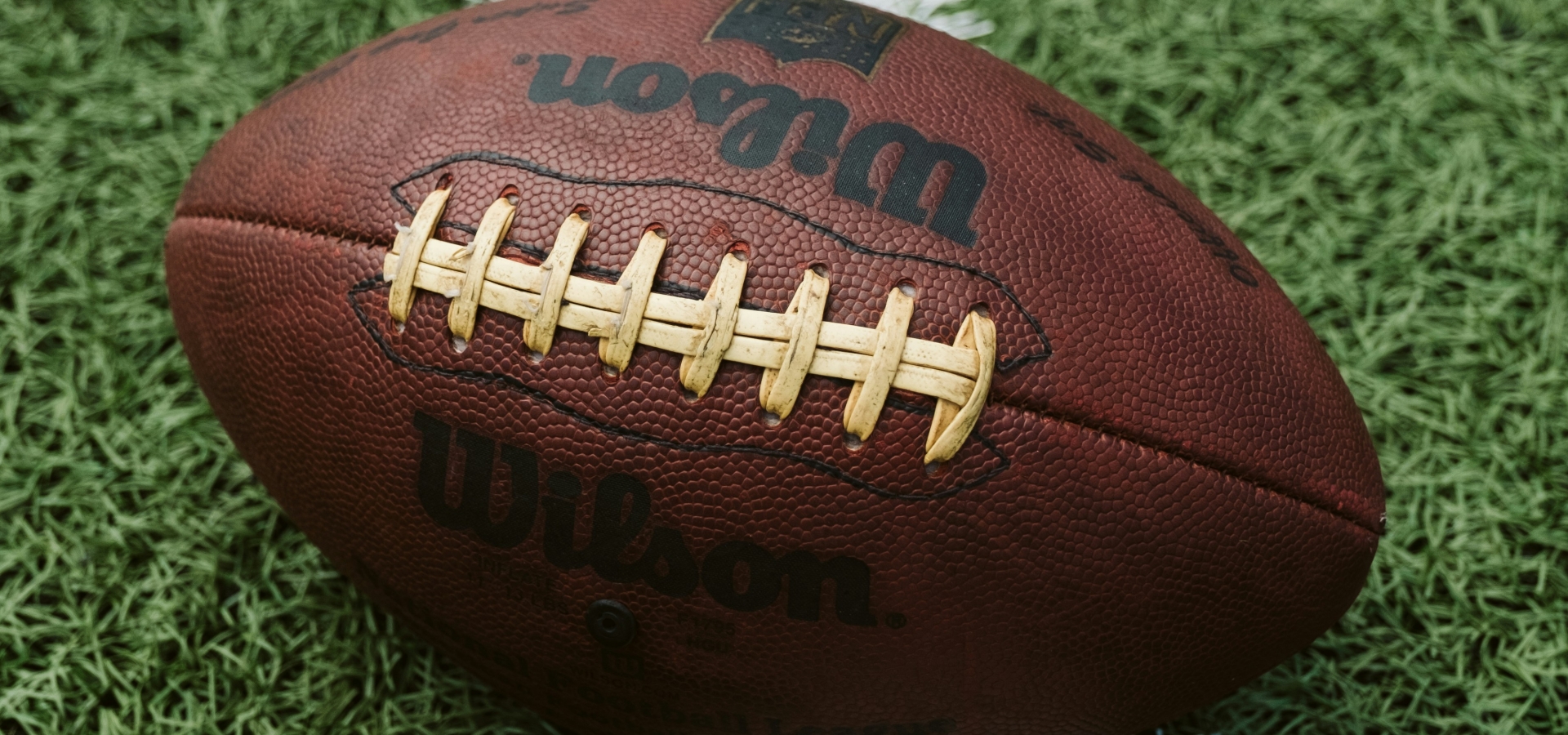 Close-up of a brown Wilson football resting on green artificial turf with a white painted line nearby.