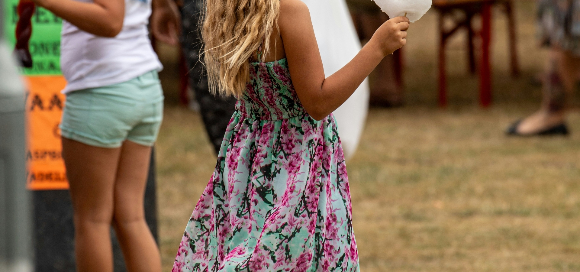 Girl in a floral dress holds white cotton candy at an outdoor festival with people in the background.