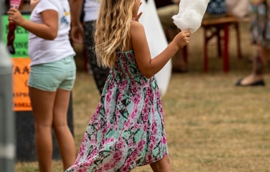 Girl in a floral dress holds white cotton candy at an outdoor festival with people in the background.