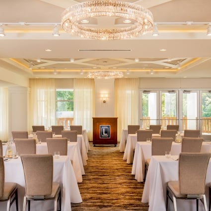 Hotel banquet room set for a conference with white tablecloths, beige chairs, and a crystal chandelier overhead.