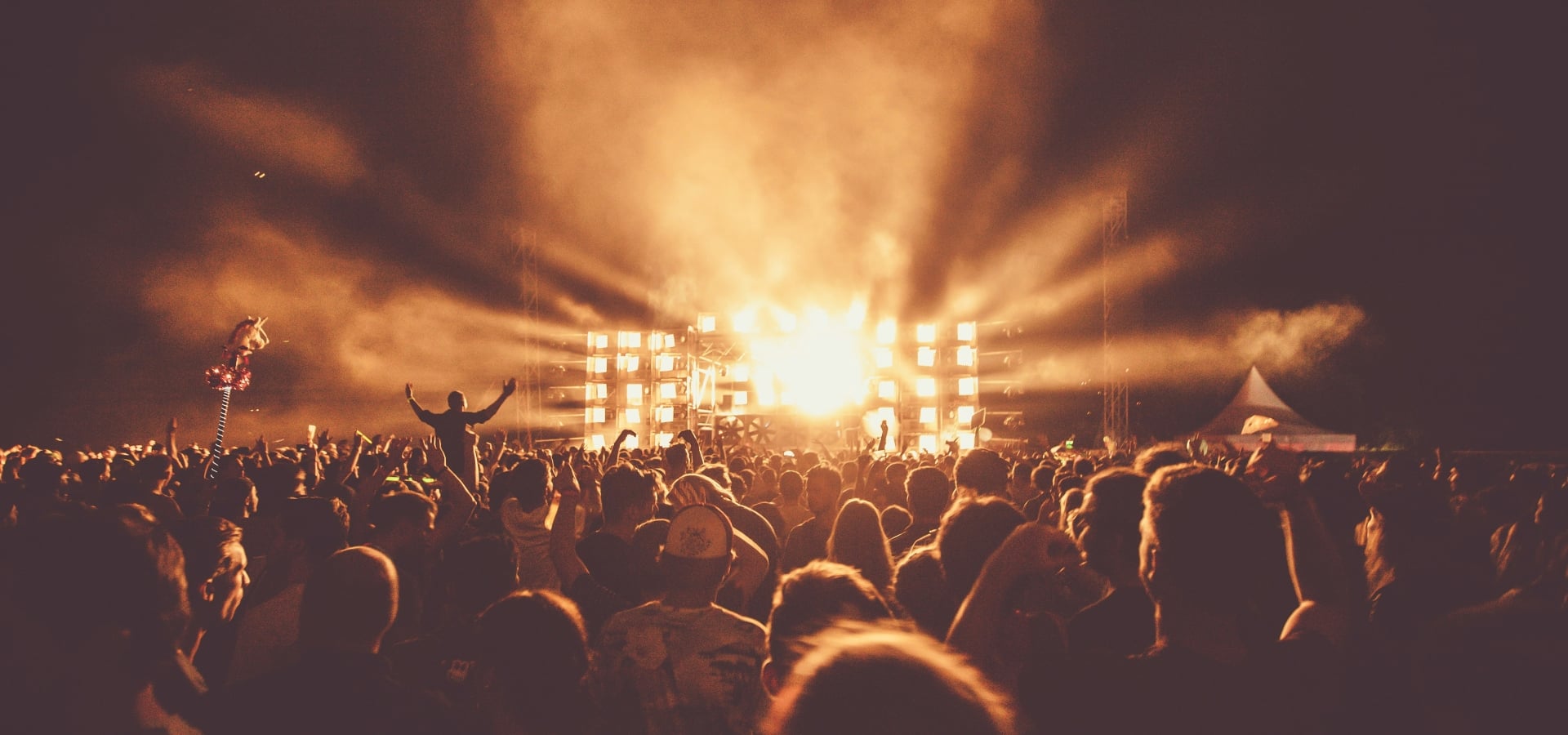 Crowd at a nighttime concert with bright stage lights and elevated performers on a lit, rectangular stage in the distance, silhouettes raised hands visible in the audience.