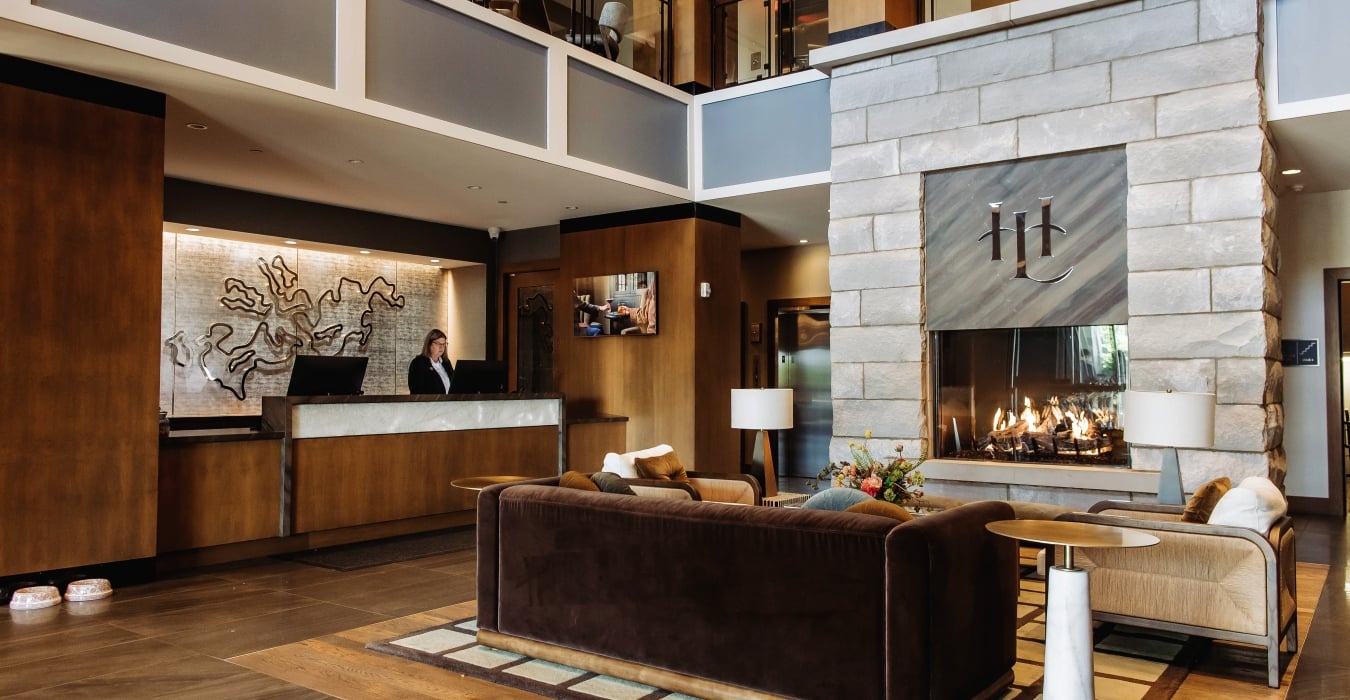 Hotel lobby scene with a dark brown reception desk, a staff member, and a large stone fireplace in a modern lounge area.