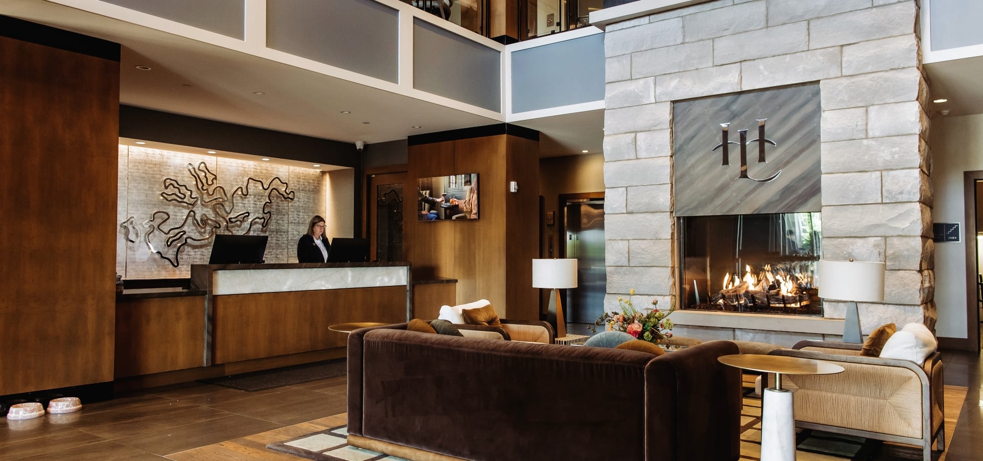 Hotel lobby scene with a dark brown reception desk, a staff member, and a large stone fireplace in a modern lounge area.