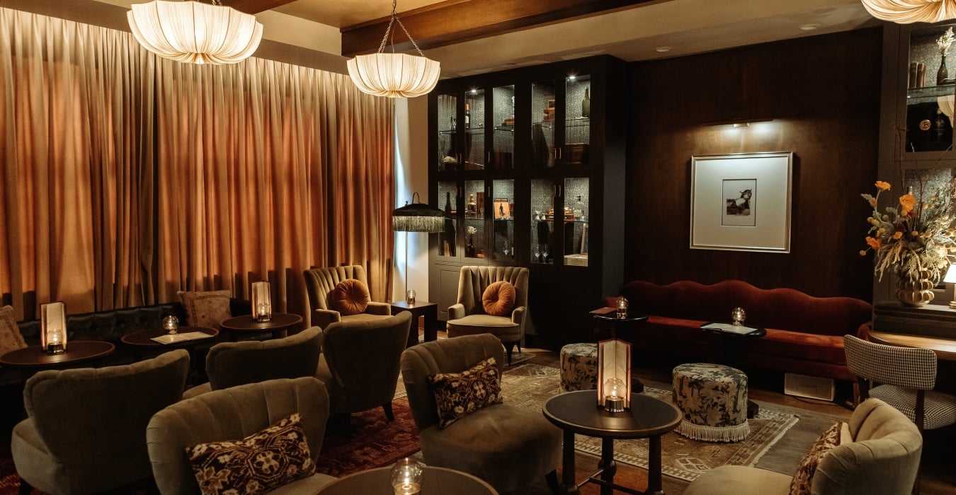 Dimly lit upscale lounge with tufted sofas, round tables, and warm pendant lights; tall curtains and a dark wood cabinet in the back.