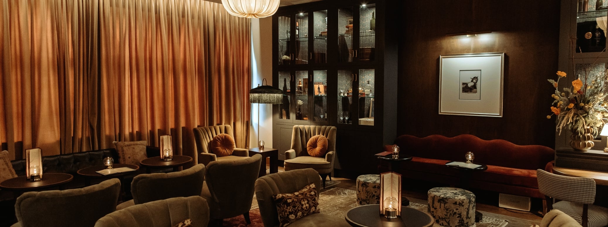 Dimly lit upscale lounge with tufted sofas, round tables, and warm pendant lights; tall curtains and a dark wood cabinet in the back.