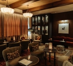 Dimly lit upscale lounge with tufted sofas, round tables, and warm pendant lights; tall curtains and a dark wood cabinet in the back.