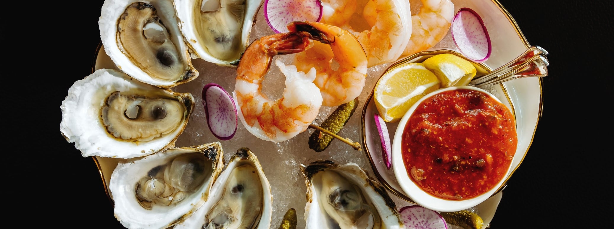 Assorted seafood platter on ice featuring oysters, shrimp, radish slices, lemon wedges, and a red dipping sauce on the side.
