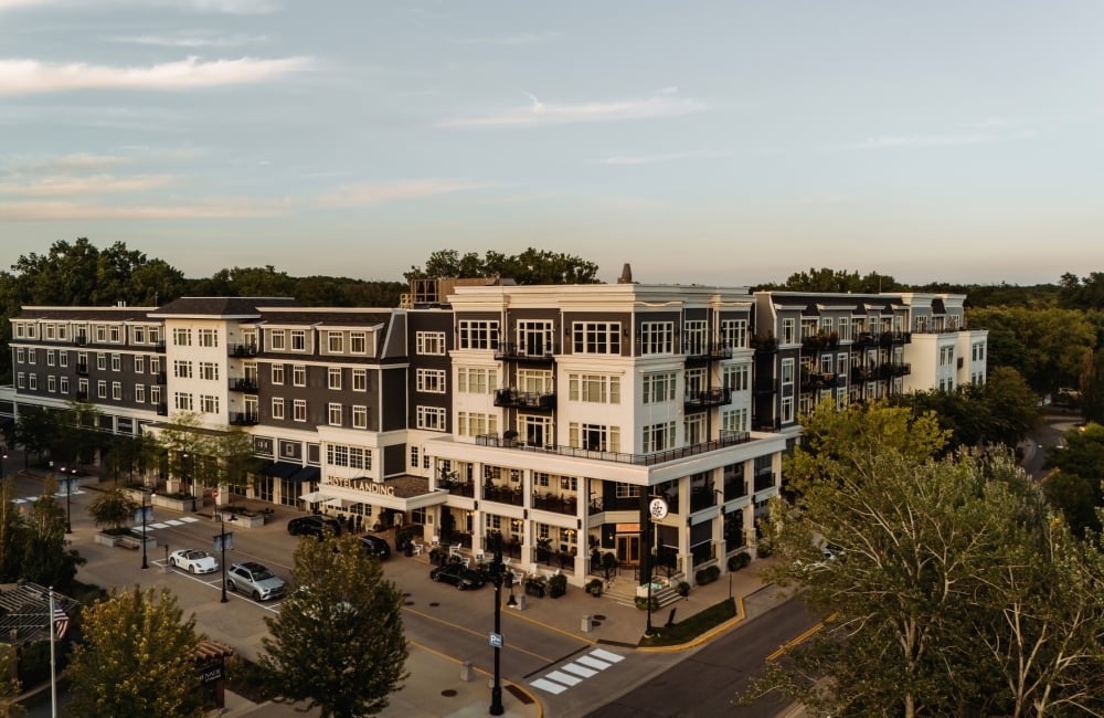 Modern multi-story residential building with balconies along a tree-lined street, storefronts on the ground floor, and cars parked nearby.
