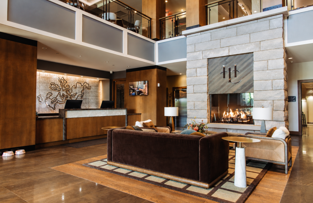 Hotel lobby with a large stone fireplace and modern seating area, brown leather sofas, and a reception desk to the left.