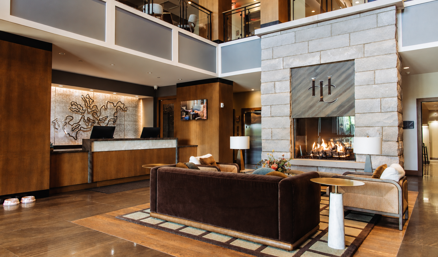 Hotel lobby with a large stone fireplace and modern seating area, brown leather sofas, and a reception desk to the left.
